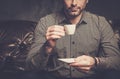 Confident handsome bearded man with cup of coffee sitting on comfortable leather sofa on dark background. Royalty Free Stock Photo