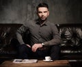 Confident handsome bearded man with cup of coffee sitting on comfortable leather sofa on dark background. Royalty Free Stock Photo