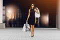 Happy, attractive young woman in a black dress and hat, wearing dark glasses, holding shopping bags in front of a shopping center Royalty Free Stock Photo