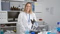 Confident, happy young blonde female scientist enjoys working, smiling while using microscope in lively lab environment