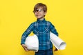 Confident little kid boy engineer or architect holding protective helmet and construction plan over yellow background. Royalty Free Stock Photo