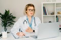 Confident mature caucasian senior female doctor in white coat using laptop for telemedicine with patients at hospital Royalty Free Stock Photo