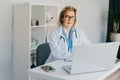 Confident mature caucasian senior female doctor in white coat using laptop for telemedicine with patients at hospital Royalty Free Stock Photo