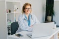 Confident mature caucasian senior female doctor in white coat using laptop for telemedicine with patients at hospital Royalty Free Stock Photo