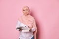 Confident Muslim woman educator in pink hijab, a school teacher holding eyeglasses and studying book, pink background