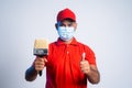 Confident painter standing with medical face mask showing thumbs up by holding paint brush while looking at camera -