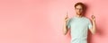 Confident and pleased young man with red hair, wearing glasses and t-shirt, pointing fingers up and smiling with smug