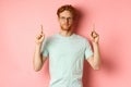 Confident and pleased young man with red hair, wearing glasses and t-shirt, pointing fingers up and smiling with smug