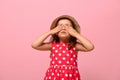 Confident portrait of gorgeous baby girl dressed in summer polka dots dress and straw hat , covering her eyes with her hands,