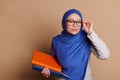 Muslim woman with head covered in blue hijab, holding books and looking at camera through eyeglasses on beige background Royalty Free Stock Photo