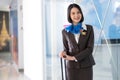 Confident pretty flight attendant/stewardess standing with luggage in front of the gate entrance. Smile and welcome on the flight