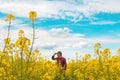 Confident and self-assured farm worker wearing red plaid shirt and trucker`s hat standing in cultivated rapeseed field in bloom