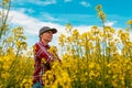 Confident and self-assured farm worker wearing red plaid shirt and trucker`s hat standing in cultivated rapeseed field in bloom