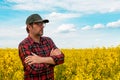 Confident and self-assured farm worker wearing red plaid shirt and trucker`s hat standing in cultivated rapeseed field in bloom