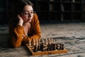 Confident sexy young woman in elegant eyeglasses thinking about chess move lying on wooden floor in dark room, looking Royalty Free Stock Photo
