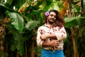 South Asian gay man with floral shirt, blue jeans stands in tropical foliage, purple flower in hair. Confident smile Royalty Free Stock Photo