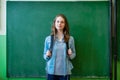 Confident smiling female high school student standing in front of chalkboard in classroom, wearing backpack, looking at camera. Royalty Free Stock Photo