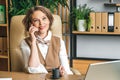 Confident stylish mature middle aged elderly woman in office.Workplace with laptop,using talking by mobile phone.General Royalty Free Stock Photo