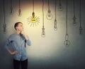 Confident teenage boy thinking, keeps one hand under chin pensive looking up choosing a hanging glowing light bulb from others are Royalty Free Stock Photo