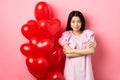 Confident teenage girl cross arms on chest and smile, celebrating valentines day in cute dress with red heart balloons Royalty Free Stock Photo