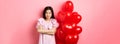 Confident teenage girl cross arms on chest and smile, celebrating valentines day in cute dress with red heart balloons Royalty Free Stock Photo