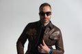 Confident unshaved guy with sunglasses fixing brown leather jacket