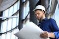 Confident young architect in navy shirt and hardhat holding a blueprint and looking at it Royalty Free Stock Photo