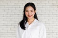 Confident young Asian woman in white shirt smiling and looking at camera on white brick background, authentic half body portrait.