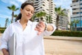 Confident young business woman checking time on her wrist watch, holding laptop, standing against modern buildings Royalty Free Stock Photo