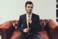 Confident young businessman straightens tie while sitting in red leather armchair .