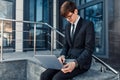 Confident young businessman, uses a laptop for work, sitting on the street against the background of a glass business center Royalty Free Stock Photo