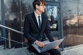 Confident young businessman, uses a laptop for work, sitting on the street against the background of a glass business center Royalty Free Stock Photo