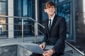 Confident young businessman, uses a laptop for work, sitting on the street against the background of a glass business center Royalty Free Stock Photo