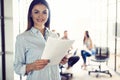 Confident young businesswoman standing with documents in a modern office with colleagues working in the background. Royalty Free Stock Photo