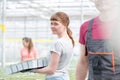 Confident young female botanist carrying seedlings by coworker in greenhouse