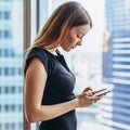 Confident young female using apps on mobile phone standing near big window in modern office Royalty Free Stock Photo