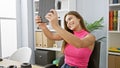 Confident young hispanic business woman grinning while working indoors; she uses her touchpad to take a beautiful office selfie Royalty Free Stock Photo