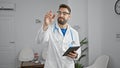 Confident young hispanic male doctor with beard in uniform, smiling and pointing finger at someone in clinic\'s waiting room, Royalty Free Stock Photo