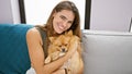 Confident young hispanic woman enjoying a fun laugh, hugging her pet dog while sitting joyfully on her home sofa with a beautiful Royalty Free Stock Photo