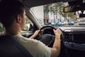 Confident young man driving car on the city streets. Teen driver keeps hands on the steering wheel looking ahead at the urban