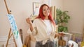 Confident young redhead woman artist holding paintbrush and palette, smiling brightly at her canvas in the heart of a bustling art Royalty Free Stock Photo