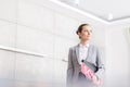 Confident young saleswoman looking away while standing with sold placard and document against wall in apartment