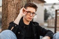 Confident young sexy man hipster straightens glasses sitting near tree on street. Fashionable portrait European guy in stylish Royalty Free Stock Photo