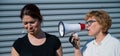 The conflict of generations. An old woman yells into a megaphone at her middle-aged daughter against gray background