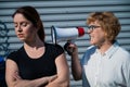 The conflict of generations. An old woman yells into a megaphone at her middle-aged daughter against gray background