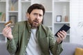 Confused adult man holding smartphone and bank card in hands while shrugging with worried expression. Uncertain customer Royalty Free Stock Photo