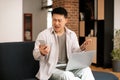 Confused asian middle aged man looking at phone screen, sitting on sofa with computer on lap, having problem with device Royalty Free Stock Photo