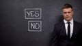 Confused businessman standing against blackboard, yes no signs drawn, hesitating