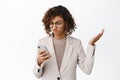 Confused corporate woman shrugging, using mobile phone with puzzled face, clueless what wrong with smartphone app