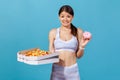 Confused doubtful woman in white sportswear holding in hands box with fatty pizza and glazed sweet donut, looking at camera with